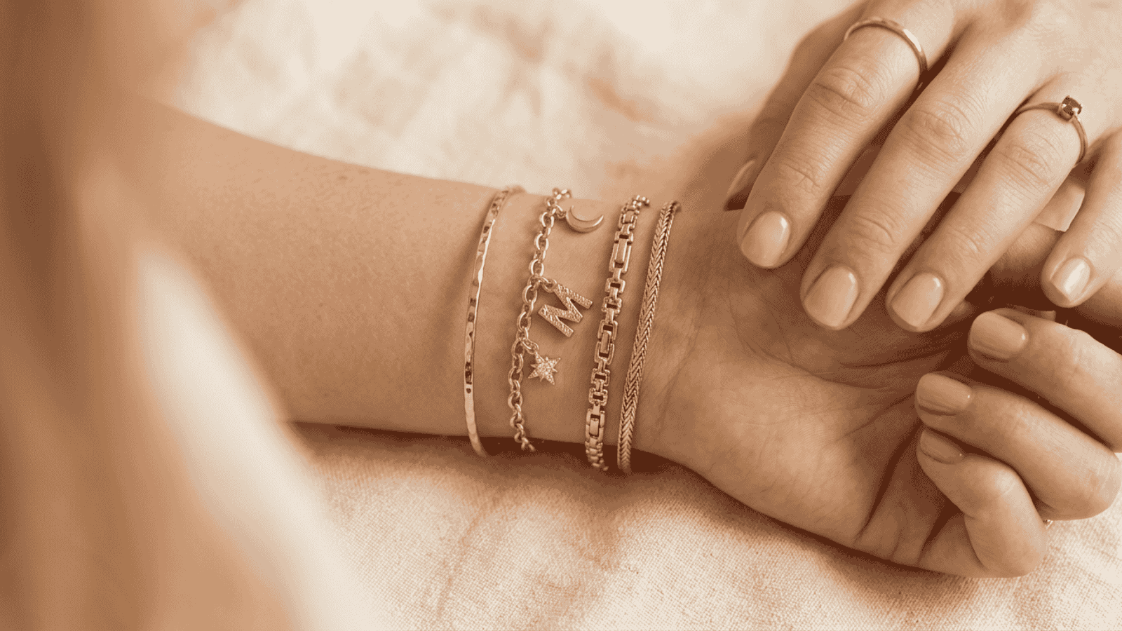 Wrist wearing a layered stack of gold plated bracelets including a chain bracelet, charm bracelet, and thin bangle on cream background