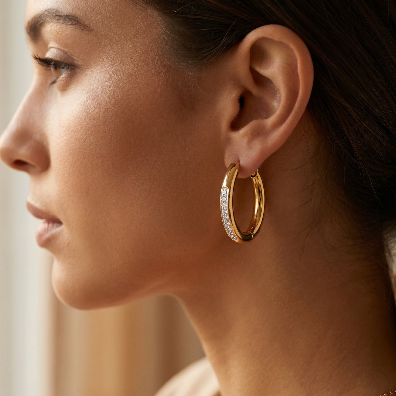 Close-up of a model's ear wearing a sculptural oversized gold plated hoop, hair tucked back in soft light