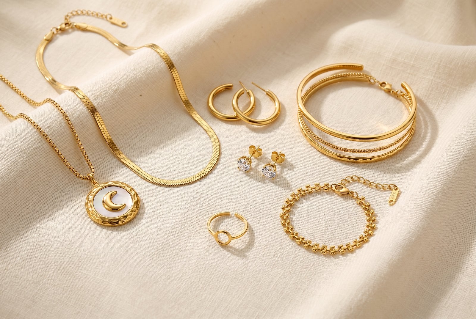 Flat-lay on cream linen of a starter set: a chain necklace, hoops, bracelet stack, ring and charm
