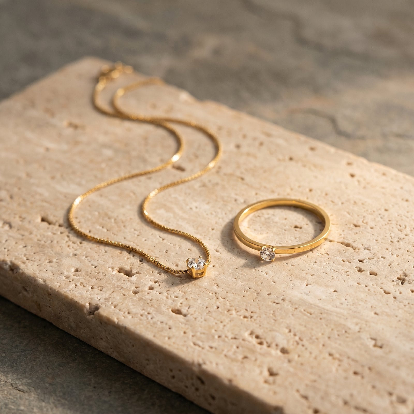 Two dainty 18K gold-plated pieces, a slim necklace and delicate ring, on warm natural stone with negative space
