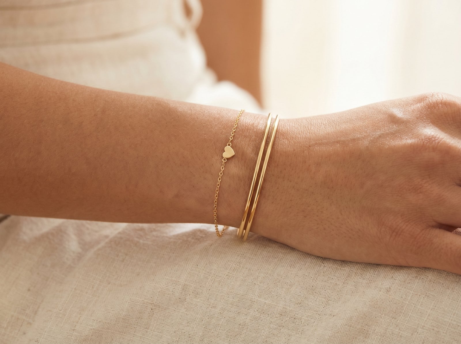 Gold heart charm bracelet stacked with two thin plain gold bangles on the wrist