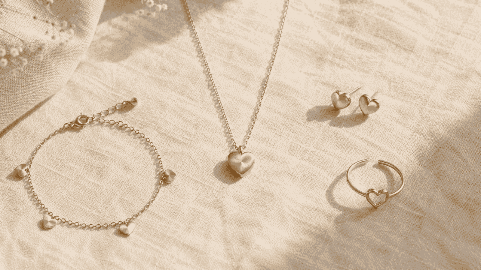 Flat lay of layered gold heart jewelry on cream linen — heart pendant necklace, heart charm bracelet, and gold stud earrings