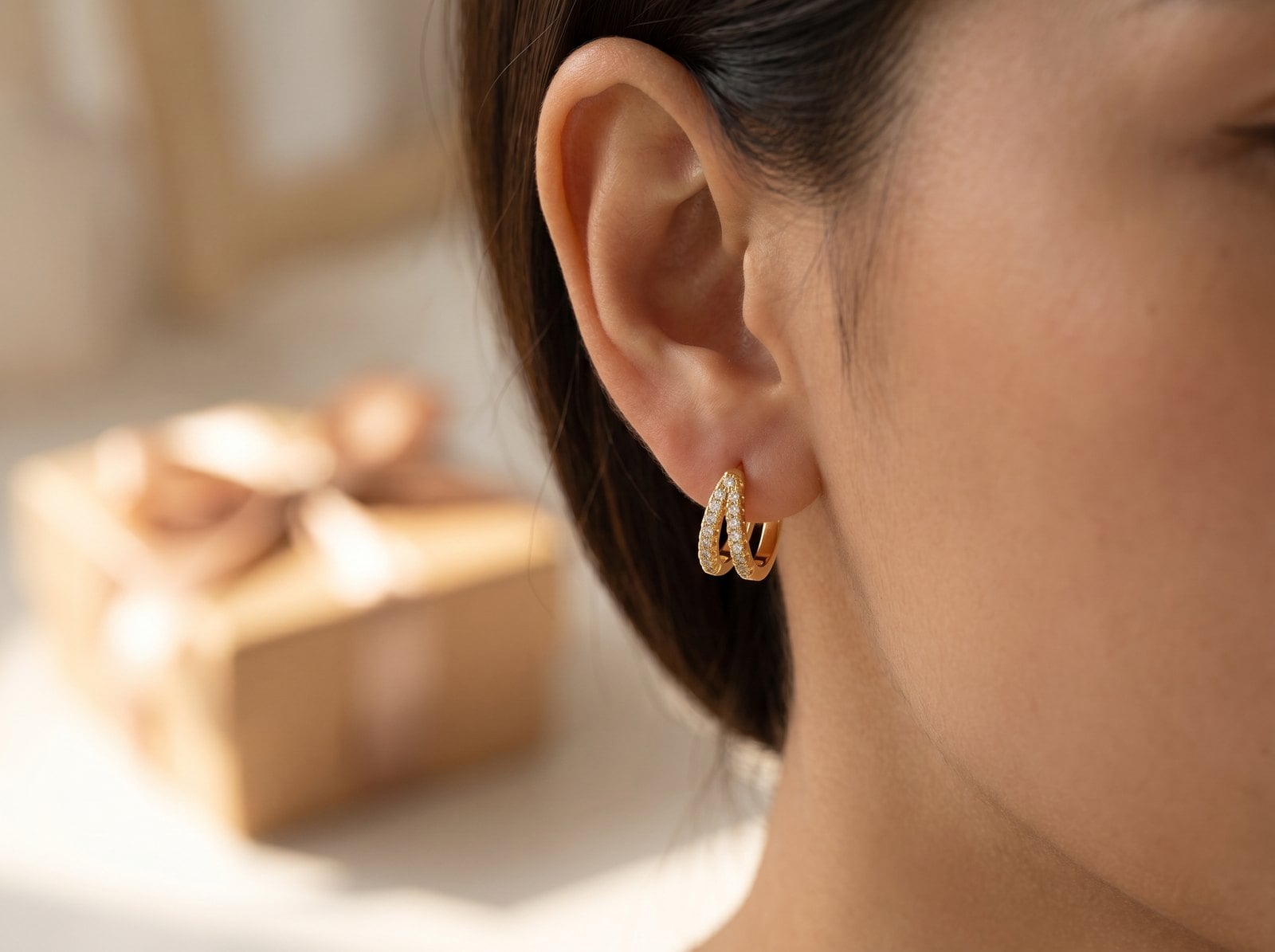 Profile crop of a woman's ear wearing a dainty gold-plated hoop, wrapped gift box blurred behind