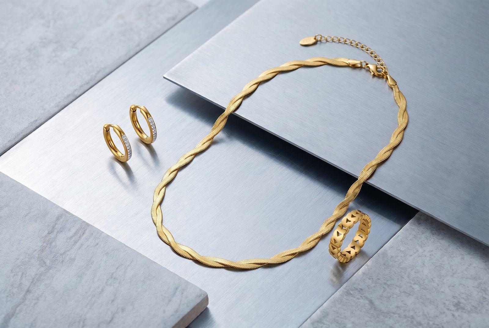 Overhead flat-lay of dainty gold-plated stainless steel pieces on a cool brushed steel surface