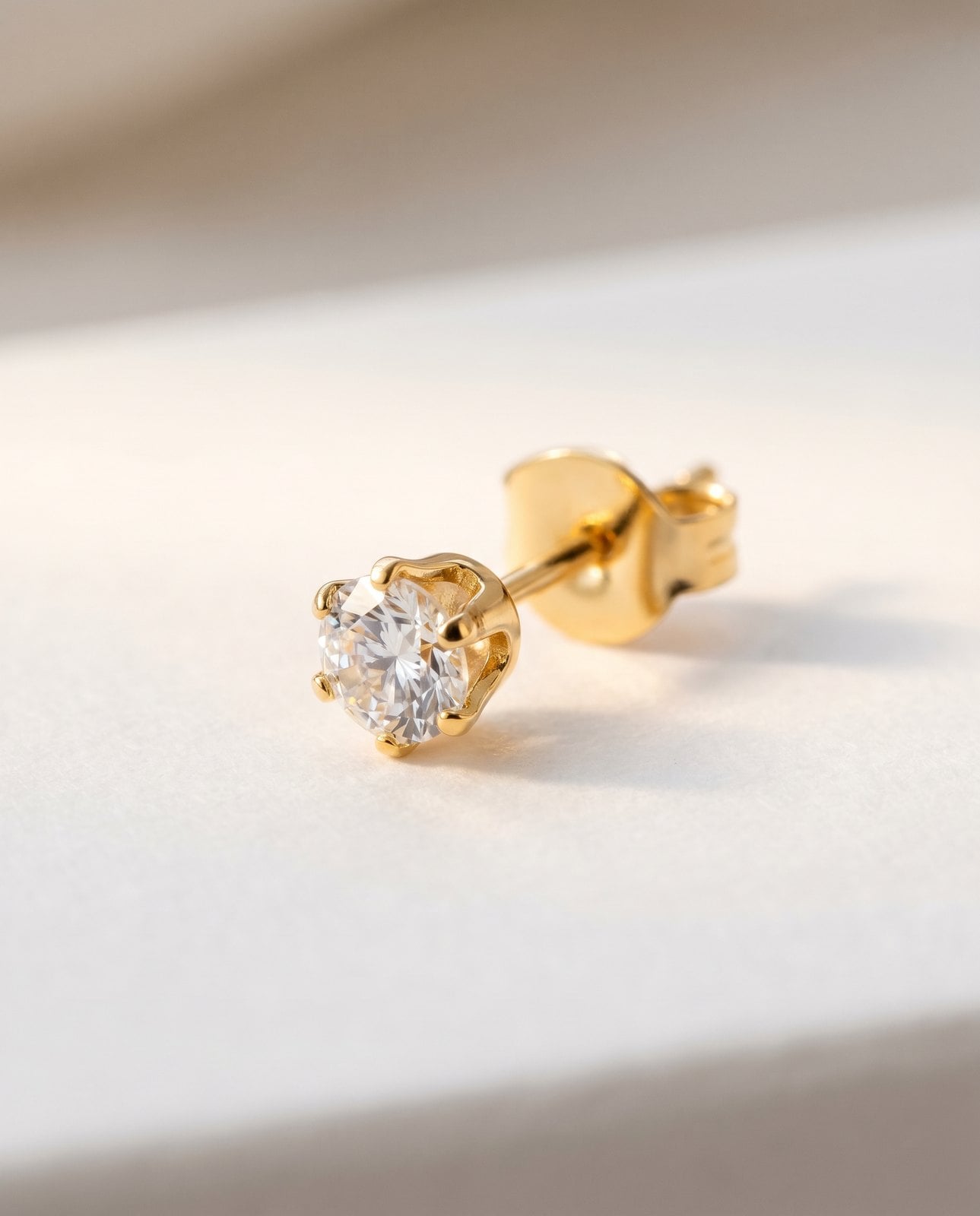 Close macro of a single dainty gold-plated stud earring on soft white showing its smooth PVD surface