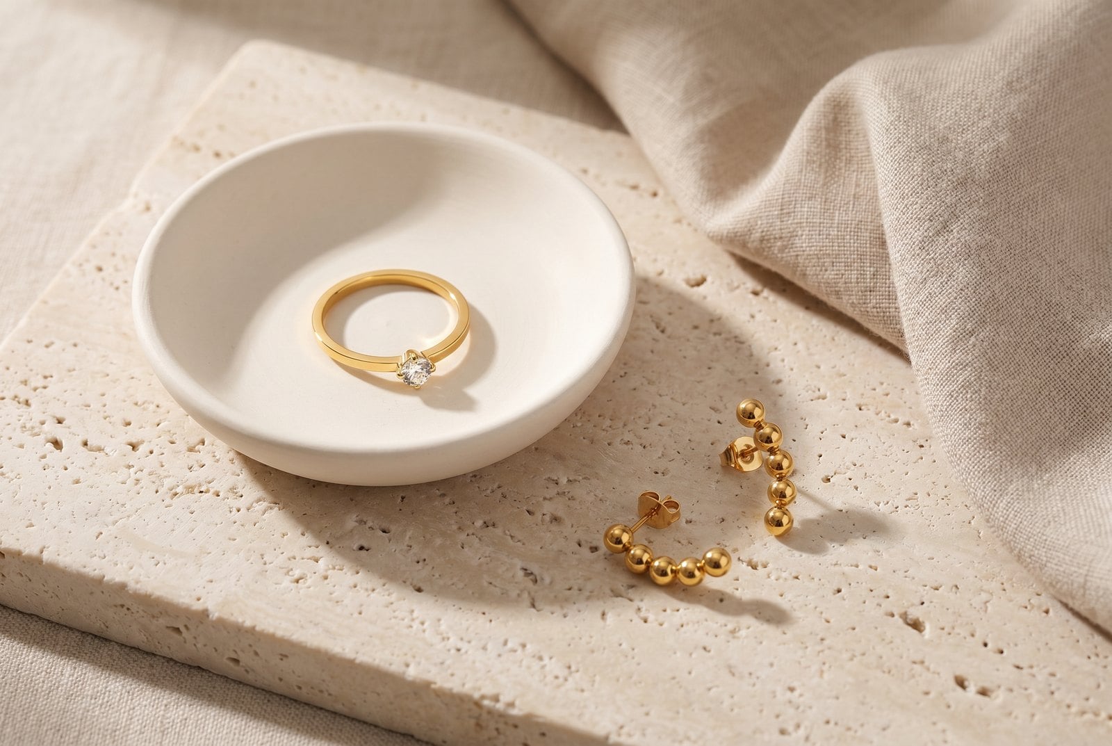 Dainty gold-plated pieces stored separately in a soft ceramic dish on pale stone with a folded cloth