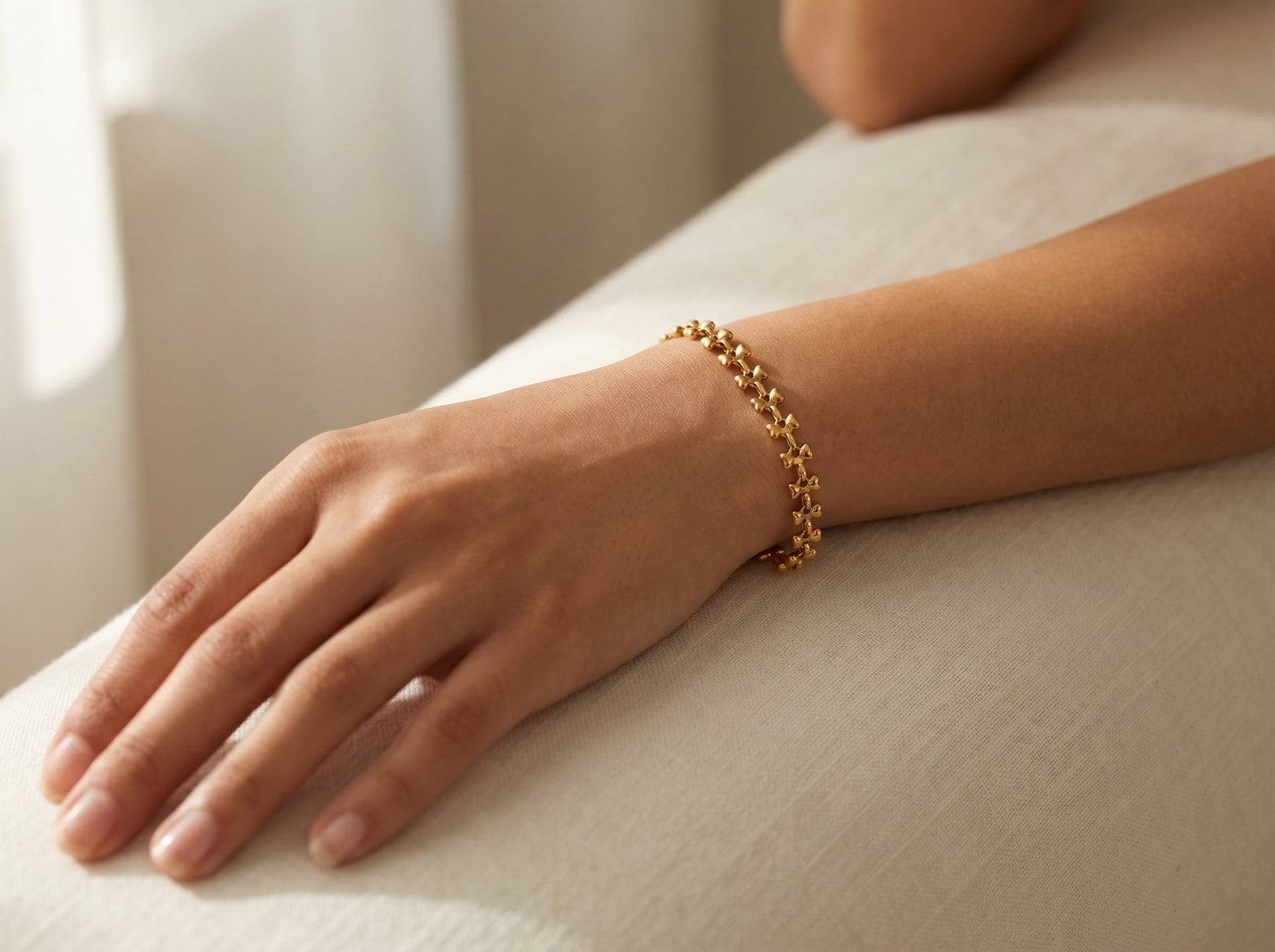 Relaxed model's wrist wearing a dainty gold-plated chain bracelet against calm skin on ivory linen