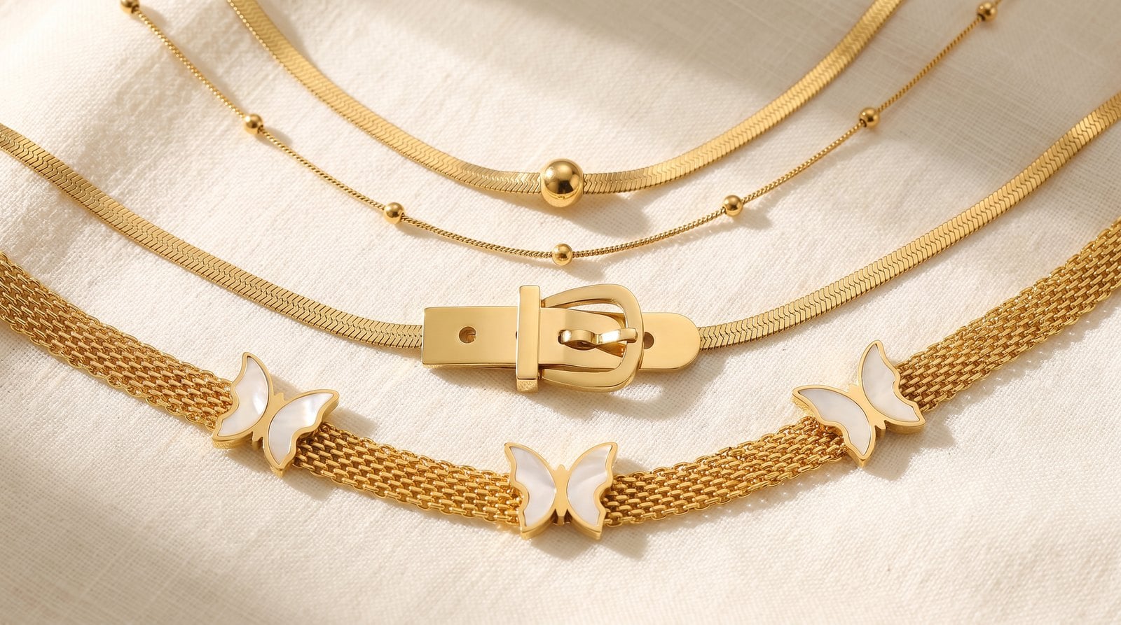 Three dainty gold-plated chain styles laid parallel on cream linen, all the same warm gold tone.