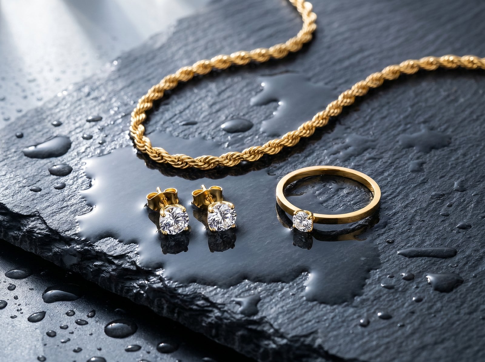 Overhead flat-lay of dainty gold-plated chain, studs and ring on wet slate with scattered water droplets.