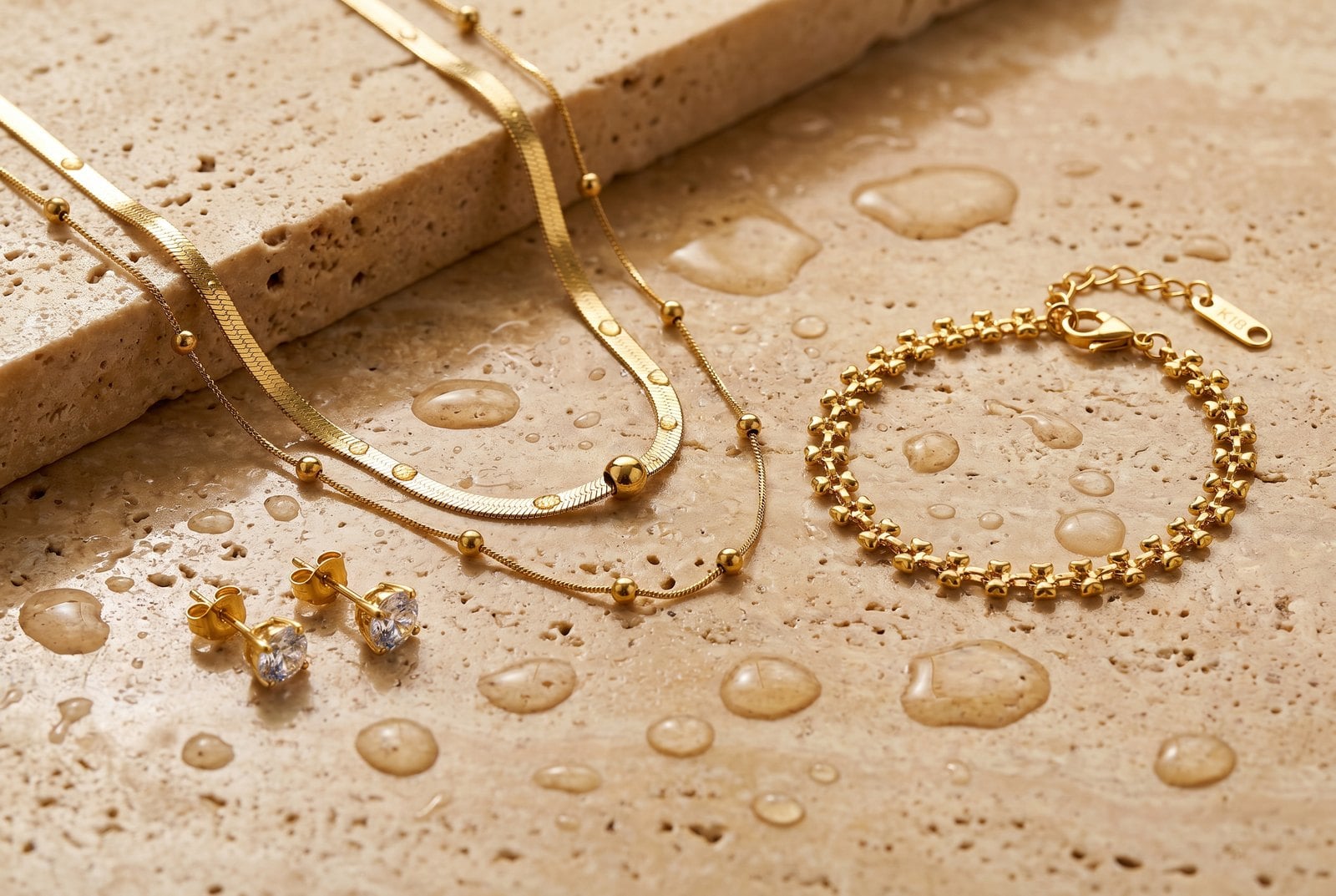 Flat-lay of 18K gold-plated necklace, bracelet and studs on warm travertine with scattered water droplets