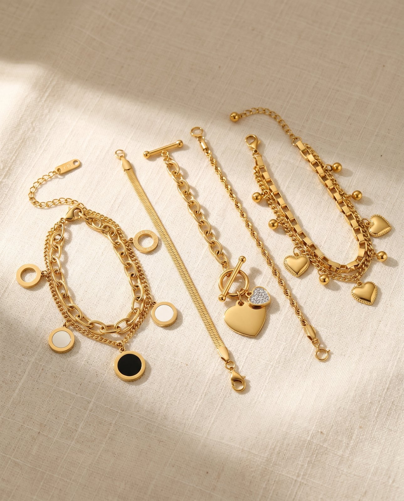 Flat-lay of five distinct gold-plated bracelets fanned on cream linen showing varied thickness and texture