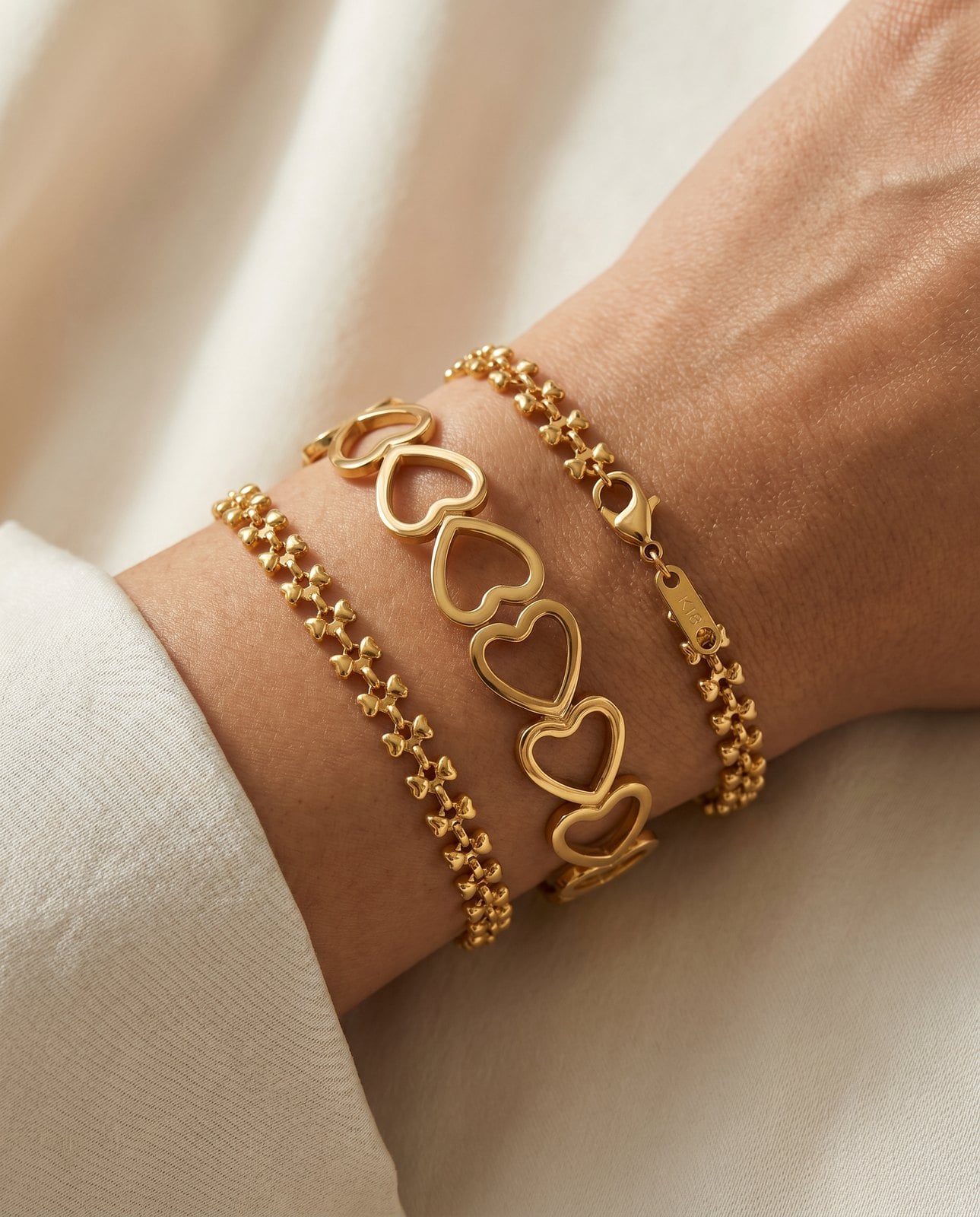 Wrist wearing one wide gold-plated statement cuff framed by two slim delicate chain bracelets on cream