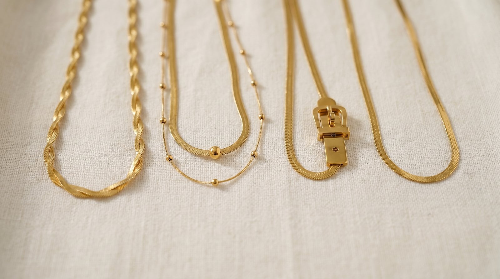 Five near-identical slim gold-plated chains lined up in a row on cream linen