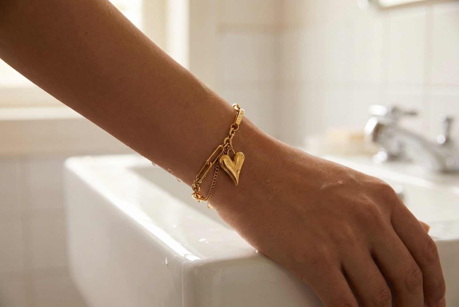 Gold-plated chain bracelet on a wrist near a sink with light water droplets on the skin