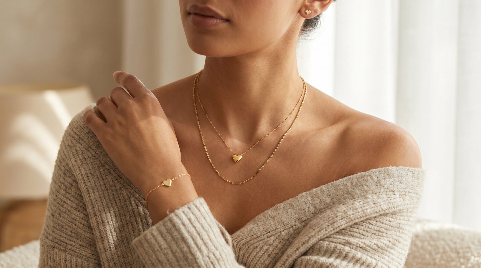 Warm-toned model wearing layered dainty gold heart jewelry — a heart pendant necklace, heart charm bracelet, and gold heart stud earrings