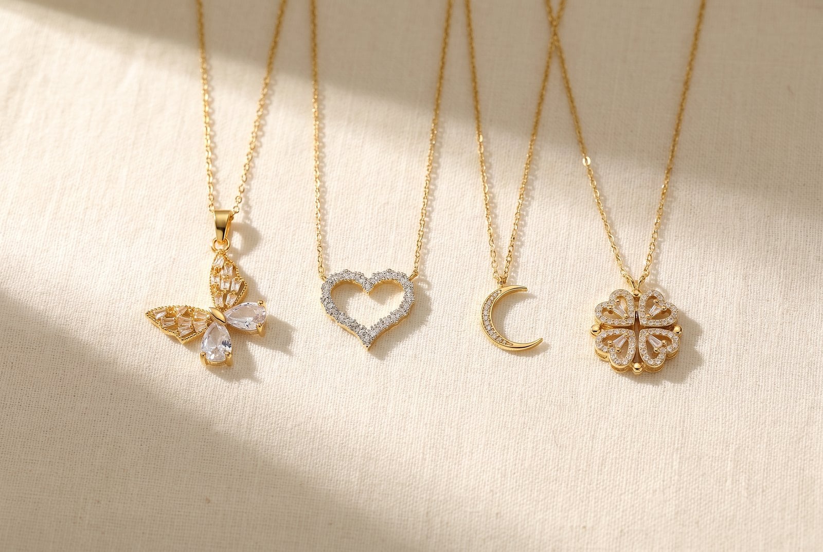 Flat-lay of four gold-plated symbol pendants — butterfly, crescent moon, heart and clover — on cream linen