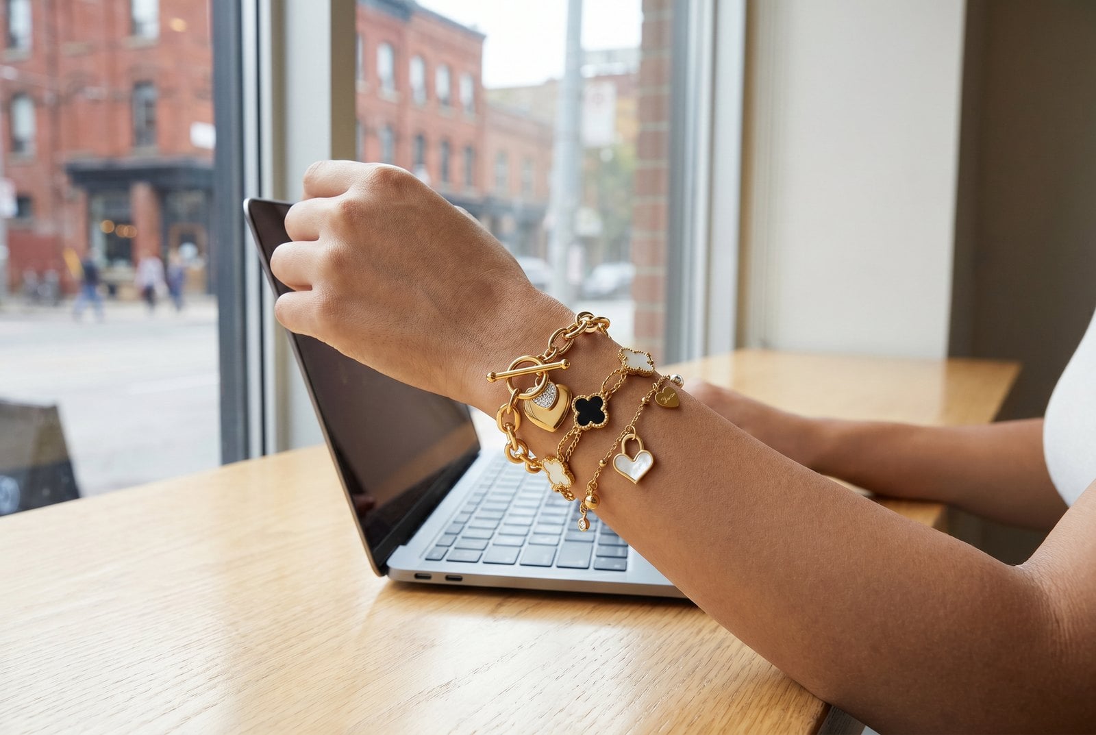 Wrist with a thin-plus-one-thick gold bracelet stack resting beside a laptop in a cafe
